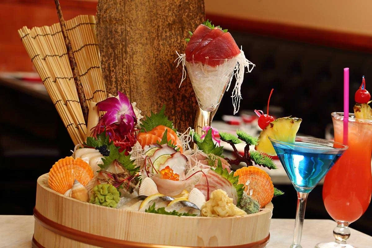 A wooden boat-shaped platter with sashimi, garnished with flowers, greenery, and shells, accompanied by colorful cocktails with fruit garnishes in a restaurant setting.