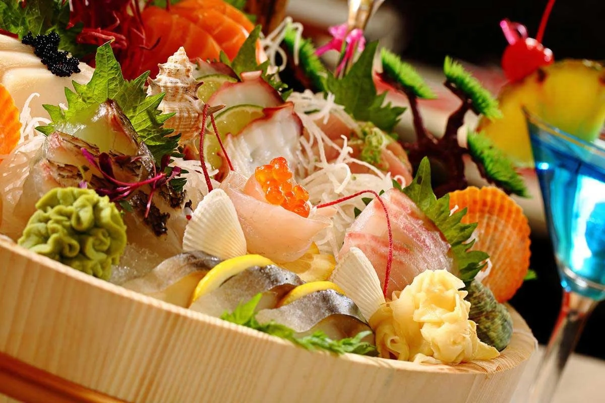 Assorted sashimi and sushi ingredients on a wooden platter, including slices of raw fish, shellfish, lemon slices, wasabi, and garnishes, with colorful vegetable decorations and a blue cocktail in the background.