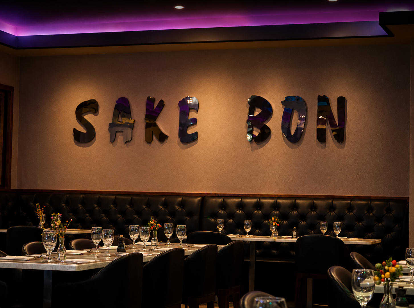 Interior of a restaurant named Shake Bon with tables set with wine glasses and small flower arrangements, and a wall with large metallic letters spelling 'SHAKE BON' under purple lighting.