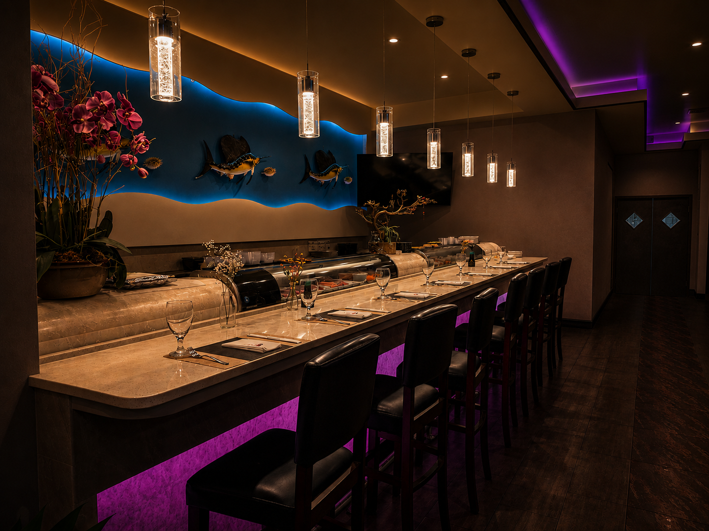 A sushi bar with a long marble counter, black leather chairs, and pendant lights hanging above. The wall features a blue background with metal fish sculptures and pink orchids in a large pot. Under the counter lighting is purple.