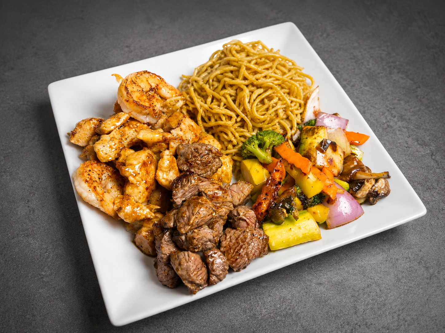 A white square plate with shrimp, beef, noodles, and a mixed vegetable stir-fry on a dark gray surface.