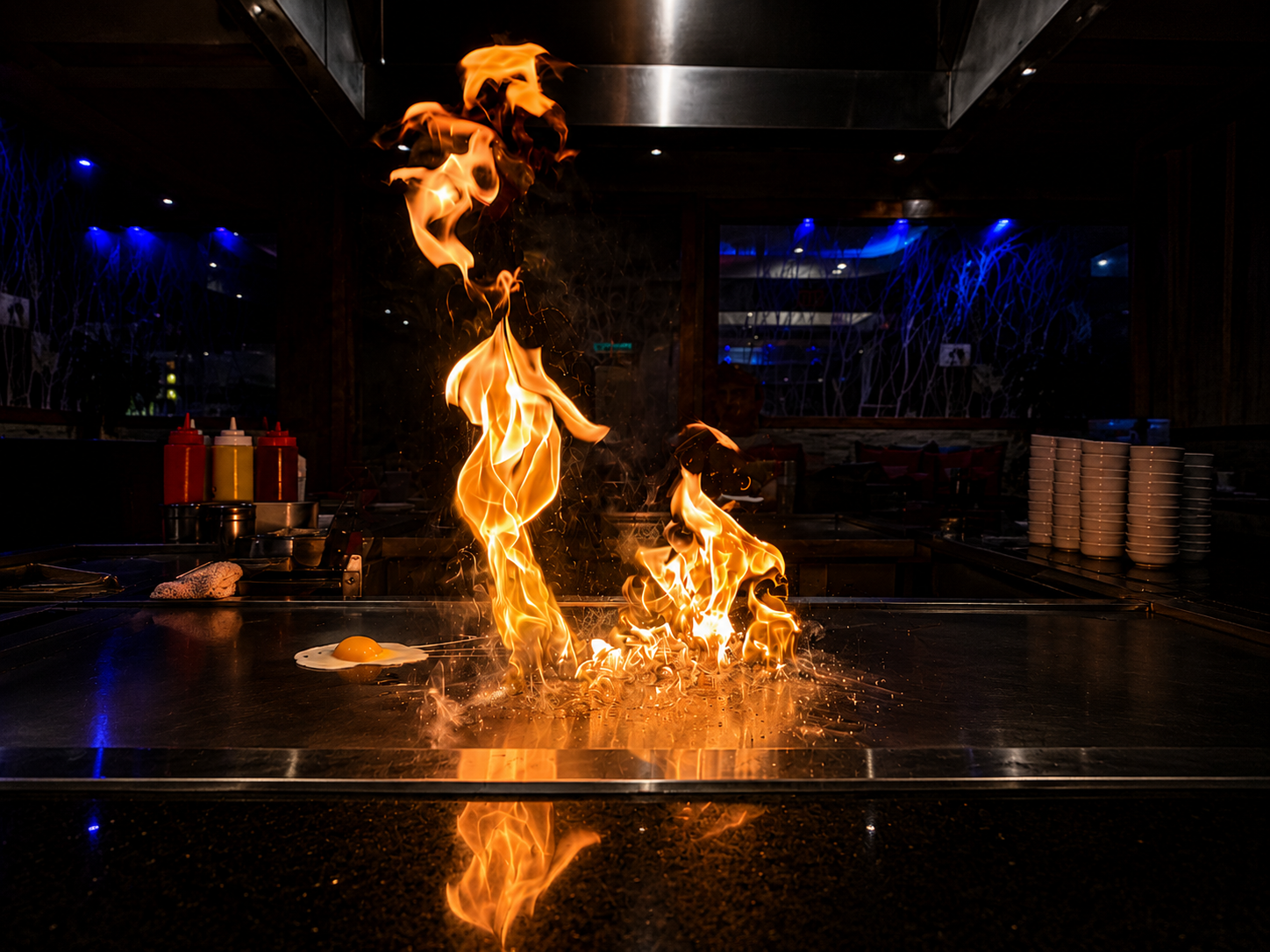 Flames and fire on a teppanyaki grill with a fried egg and ketchup bottles in the background, in a dimly lit restaurant.