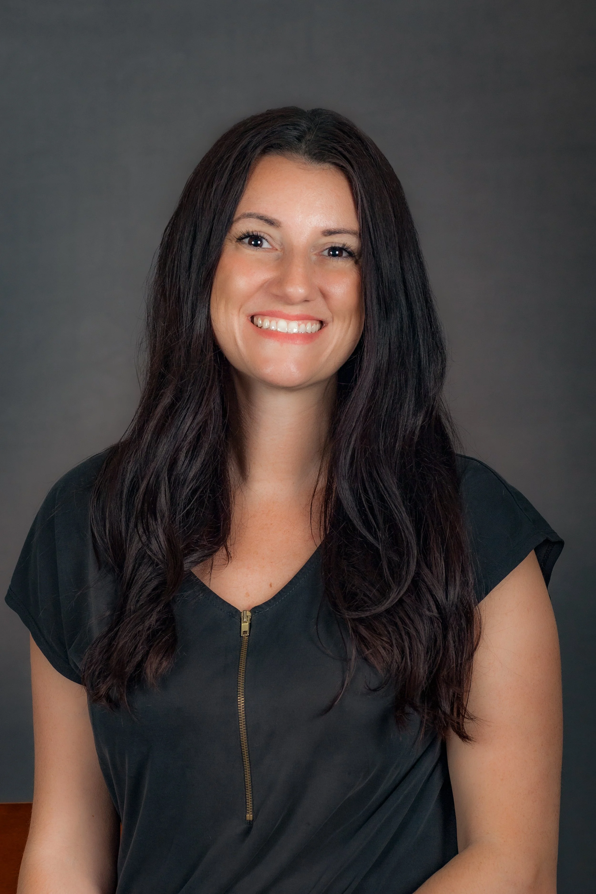 A woman with long wavy dark hair smiling, wearing a black top with a zipper in front, against a dark grey background.