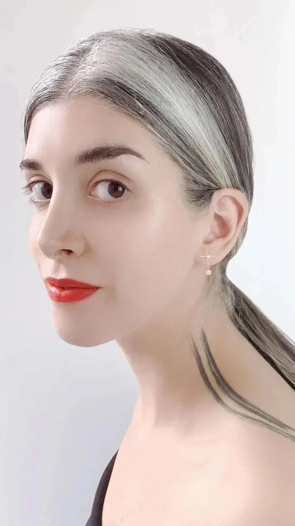 A woman with gray and black hair tied back, wearing red lipstick, star-shaped and pearl earrings, and a black top, with a tattoo on her neck, against a plain white background.