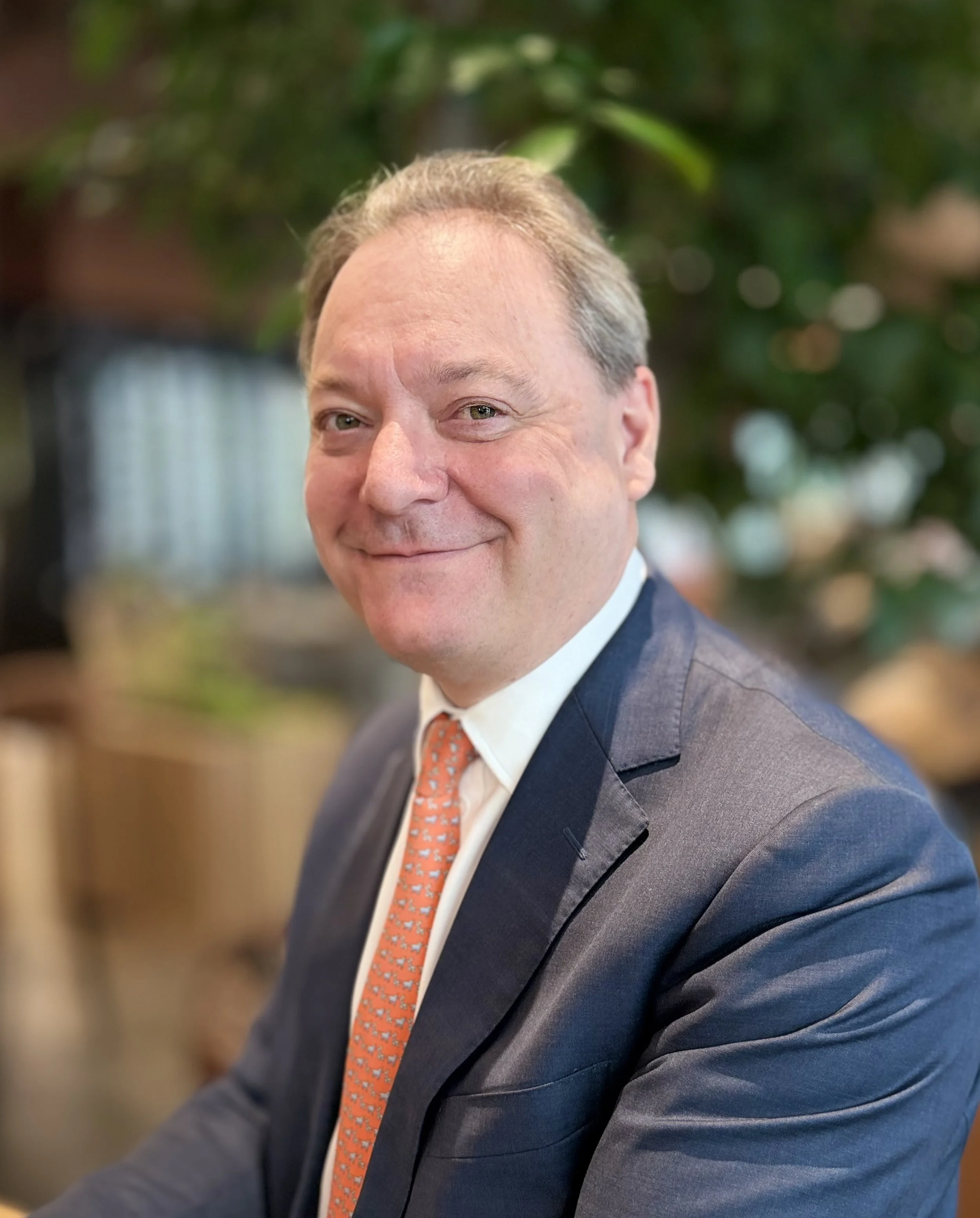 Close-up of a middle-aged man smiling, wearing a dark suit, white shirt, and orange patterned tie, with a blurred background of green plants.