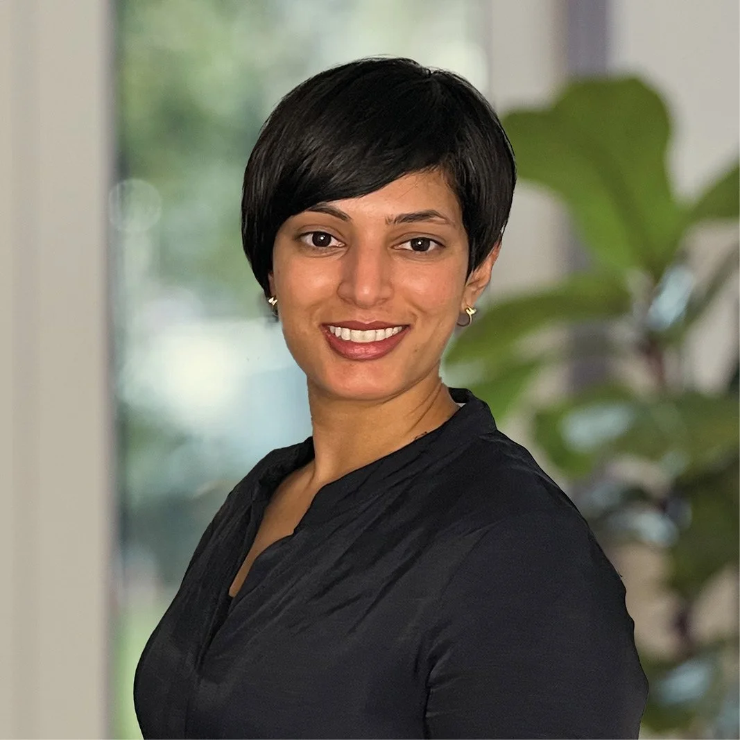 A woman with short black hair, smiling, wearing a black top, standing indoors with a large plant and window in the background.