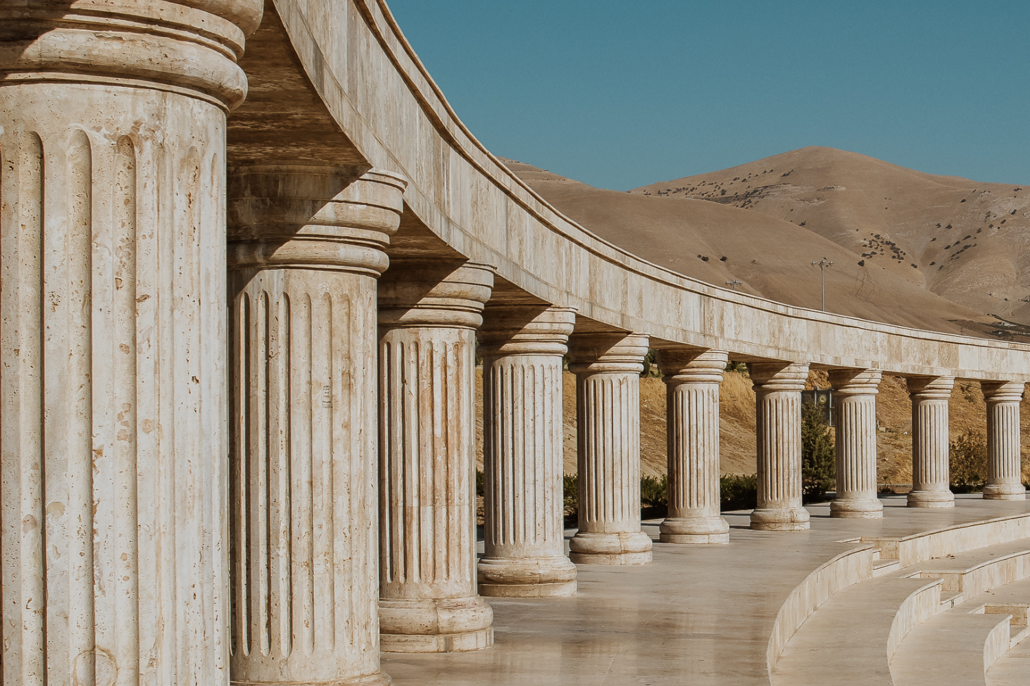 A row of classical stone columns supporting a curved structure, with dry hills in the background under a clear blue sky.