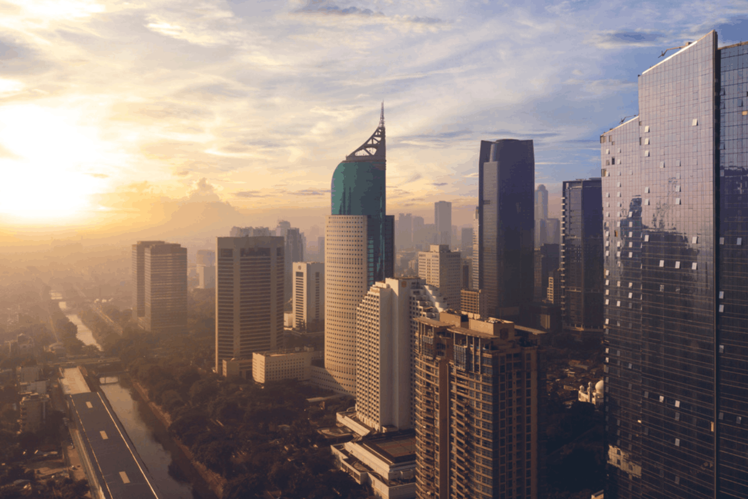 A city skyline during sunset featuring tall skyscrapers, including one with a distinctive curved top, reflecting the sunset light, with a river and a road running alongside it.