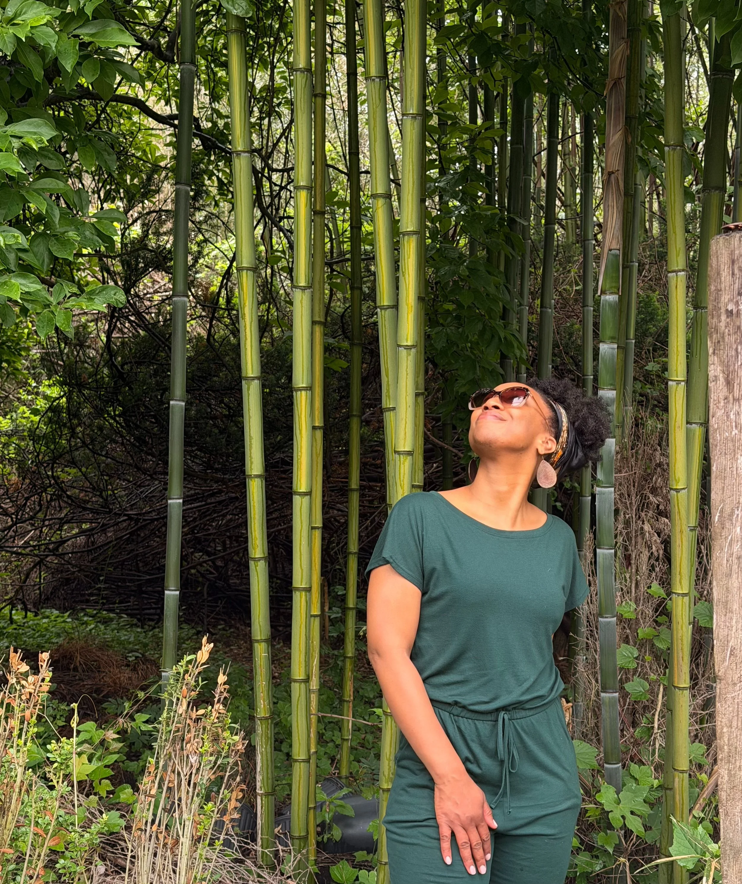 A woman with sunglasses and earrings standing in front of a bamboo grove and greenery, smiling and relaxed.