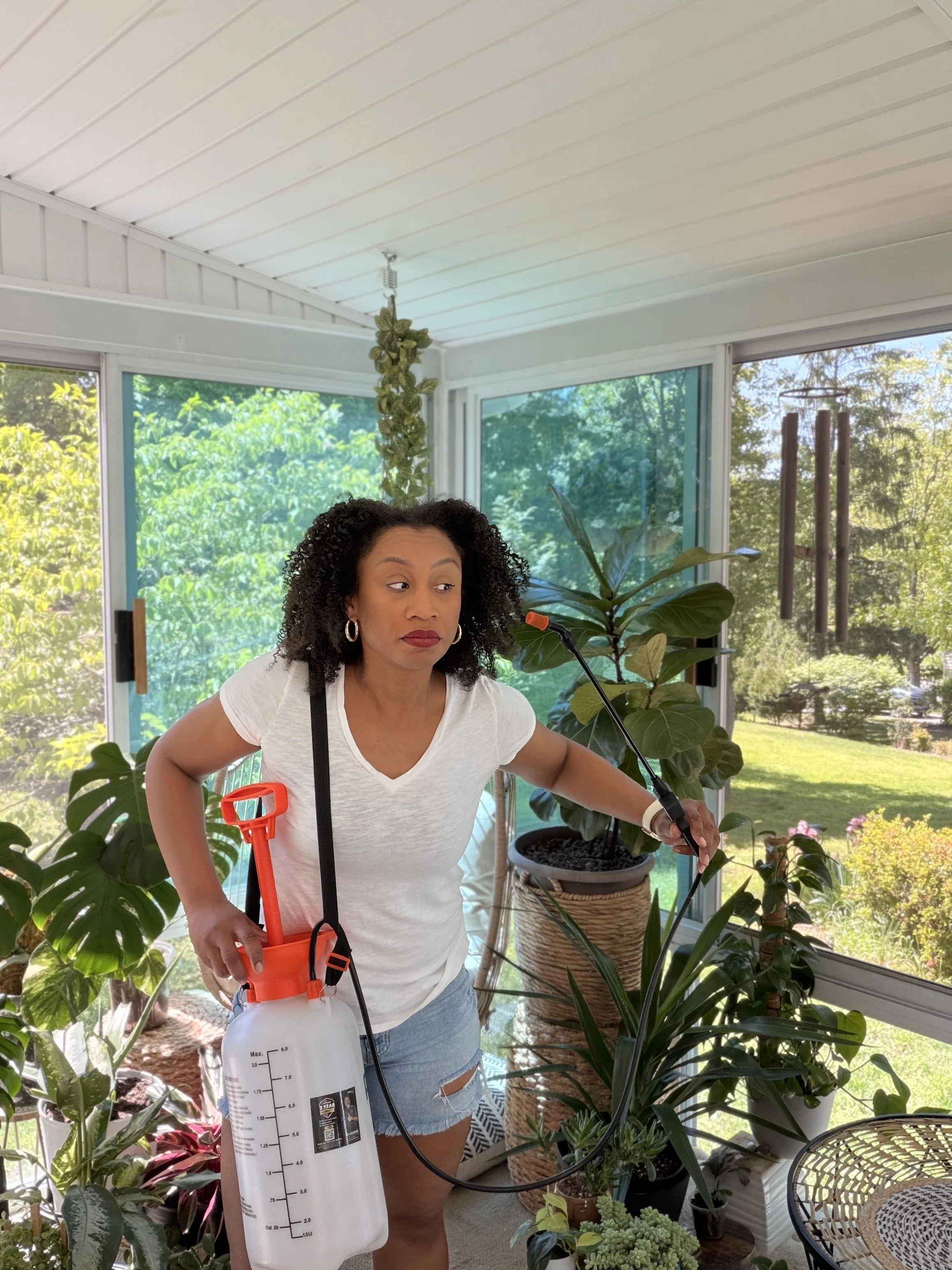 A woman spraying plants inside a greenhouse, holding a white spray bottle. She has curly black hair, is wearing a white T-shirt and ripped shorts, with a red strap around her shoulder. There are various potted plants around her, with large windows showing greenery outside.