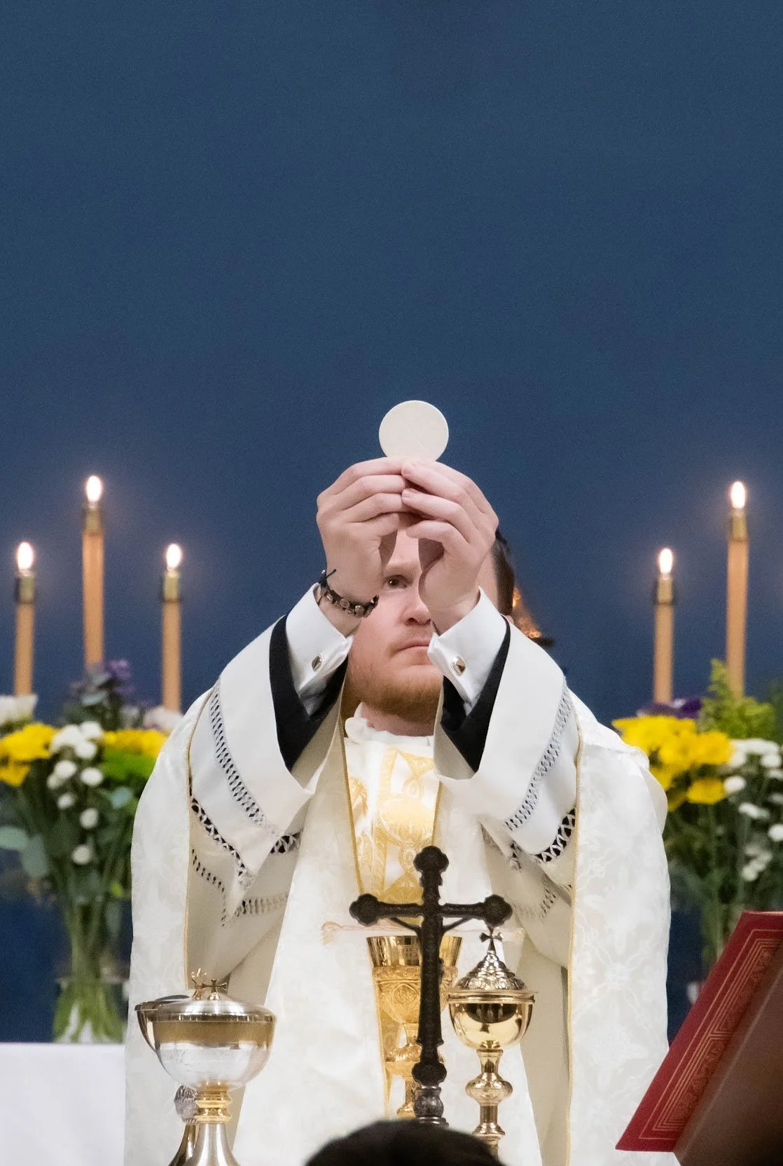 A man priest wearing white liturgical vestments holds up a communion wafer during a church service, with candles and flowers in the background.