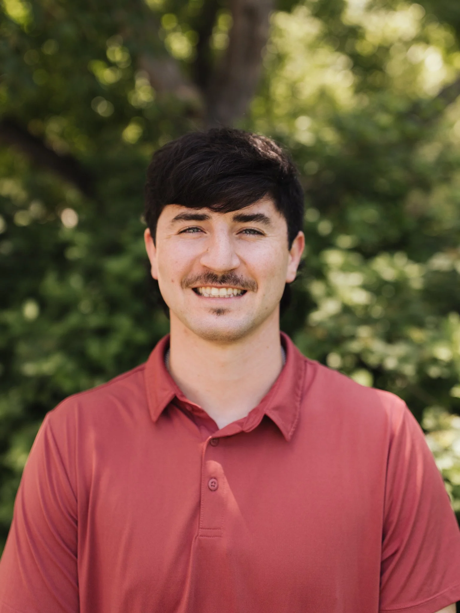 A young man with dark hair and facial hair smiling outdoors in front of green trees, wearing a reddish-pink collared shirt.