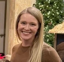 A smiling young woman with long blonde hair, standing indoors near a decorated Christmas tree.