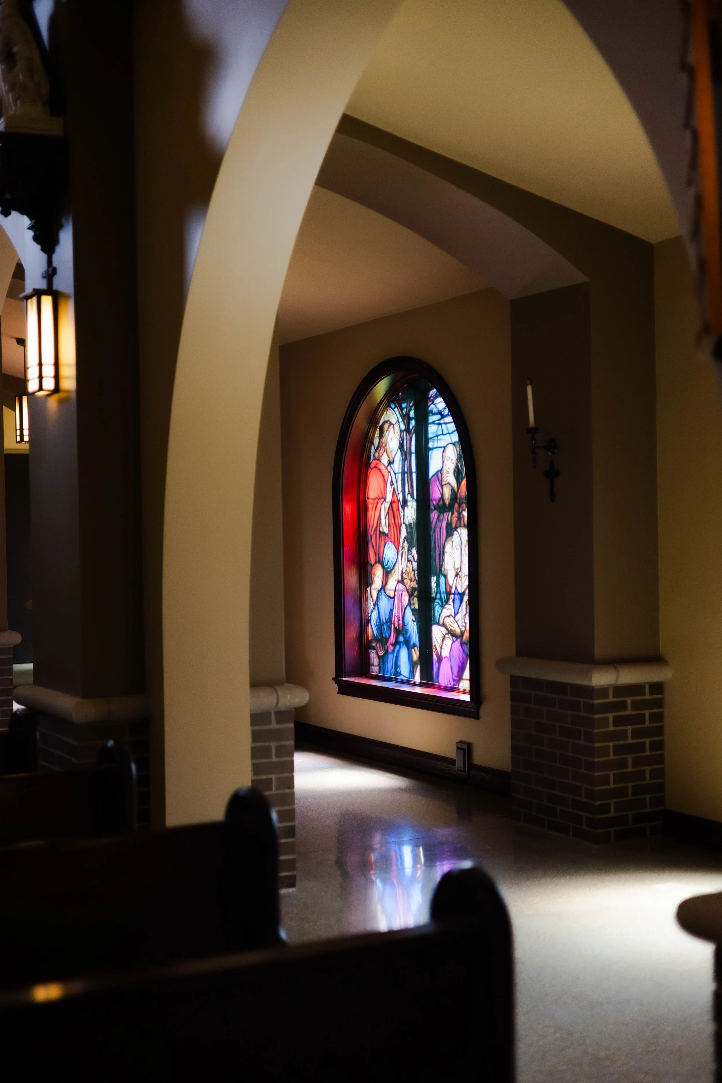 Interior of a church or chapel with a carved archway, stained glass window depicting religious figures, and wall-mounted candle holders.