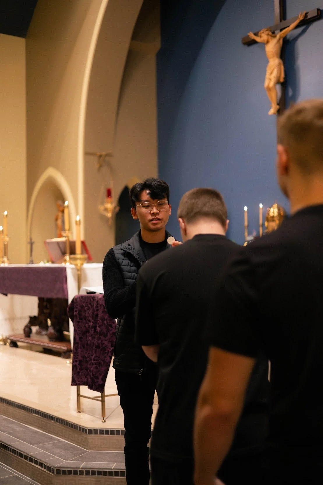 Three young men are standing and talking inside a church near an altar with religious icons, candles, and a crucifix on the wall.