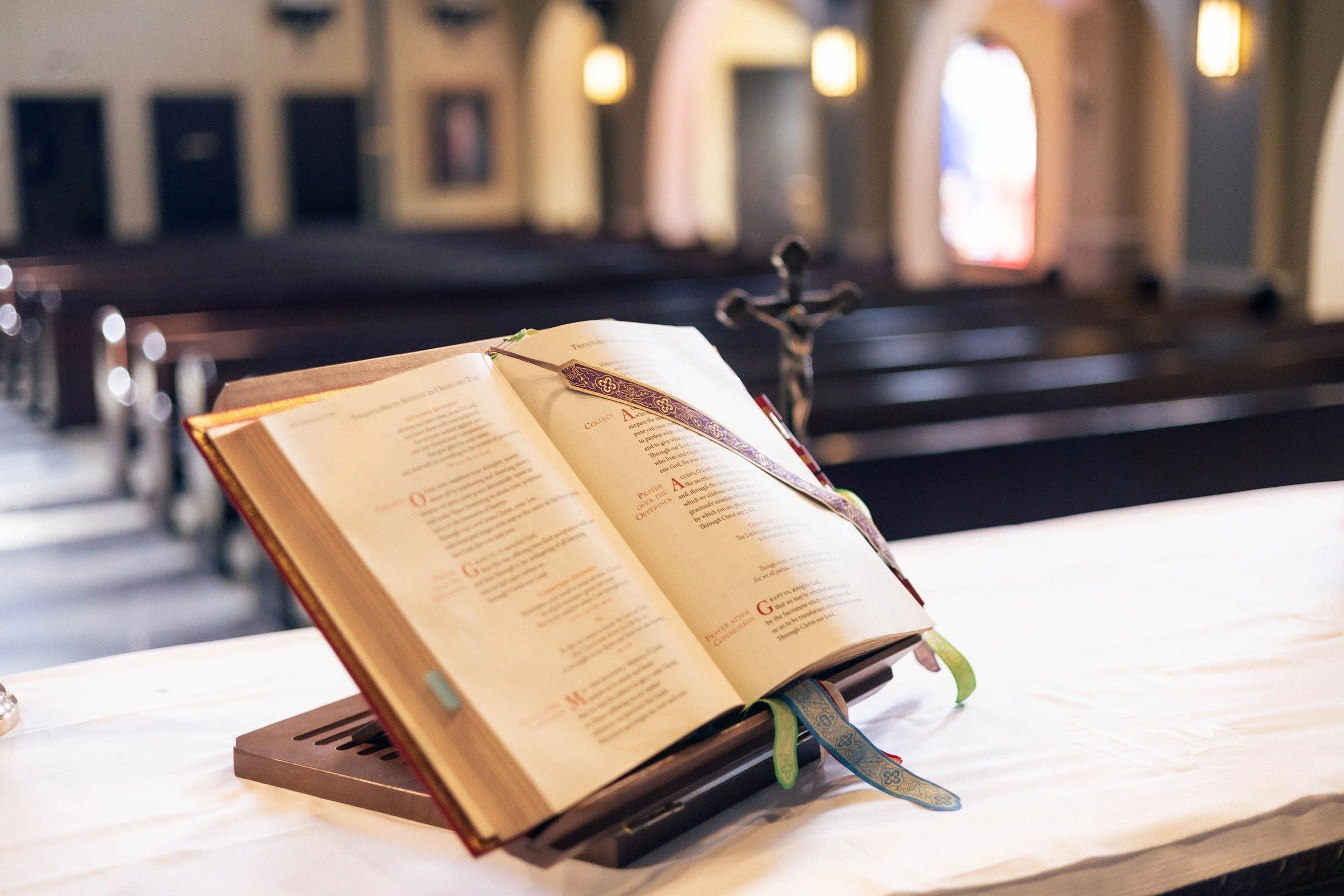 An open Bible with colorful ribbons marks on a music stand inside a church, with blurred pews and religious artwork in the background.