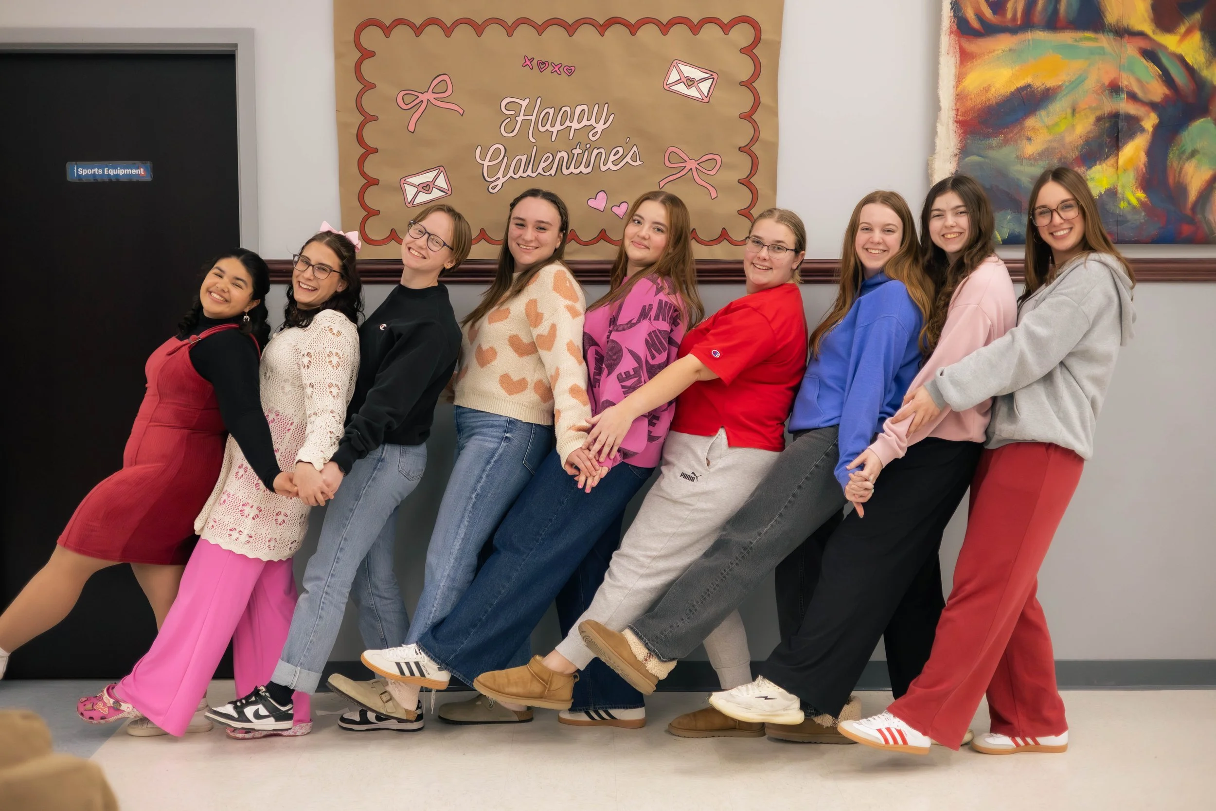 Group of nine girls standing side by side inside a building, all smiling, wearing casual clothing and colorful loungewear, with a backdrop of Valentine's Day themed decorations and artwork on the wall.