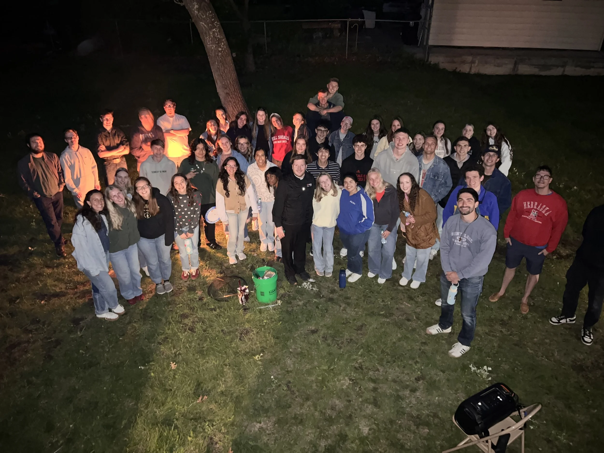 A large group of young people gathered outdoors at night, posing for a photo on a grassy area with trees and a house in the background.