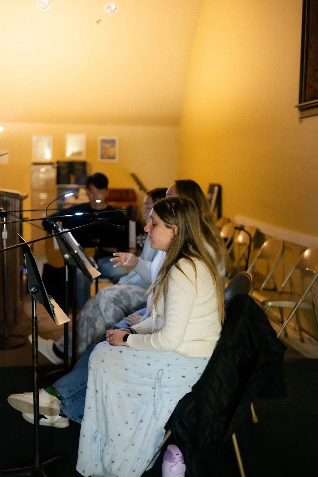 Group of people sitting in a room with music stands, some playing instruments, during Source & Summit Worship Night.