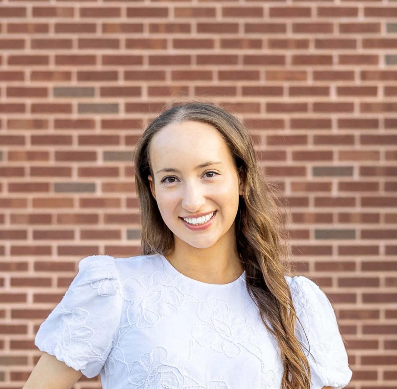 A young woman with long brown hair and a bright smile, wearing a white embroidered top, standing in front of a brick wall.
