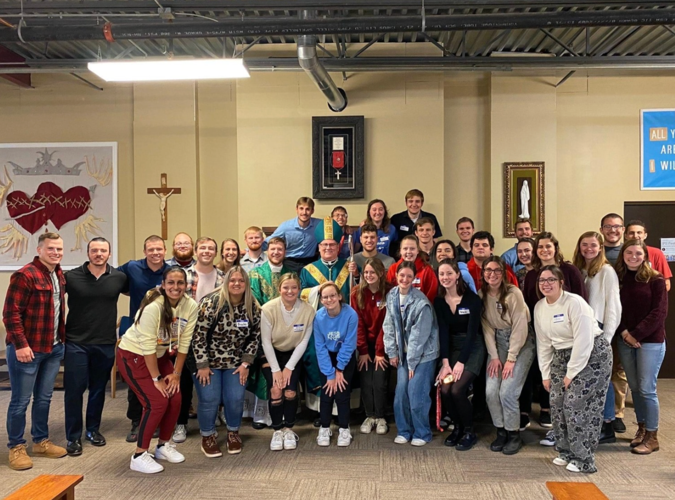 A group of people, including an individual dressed as a bishop or pope, posing for a photo in a religious setting with Christian symbols on the wall, including a crucifix and religious artwork.