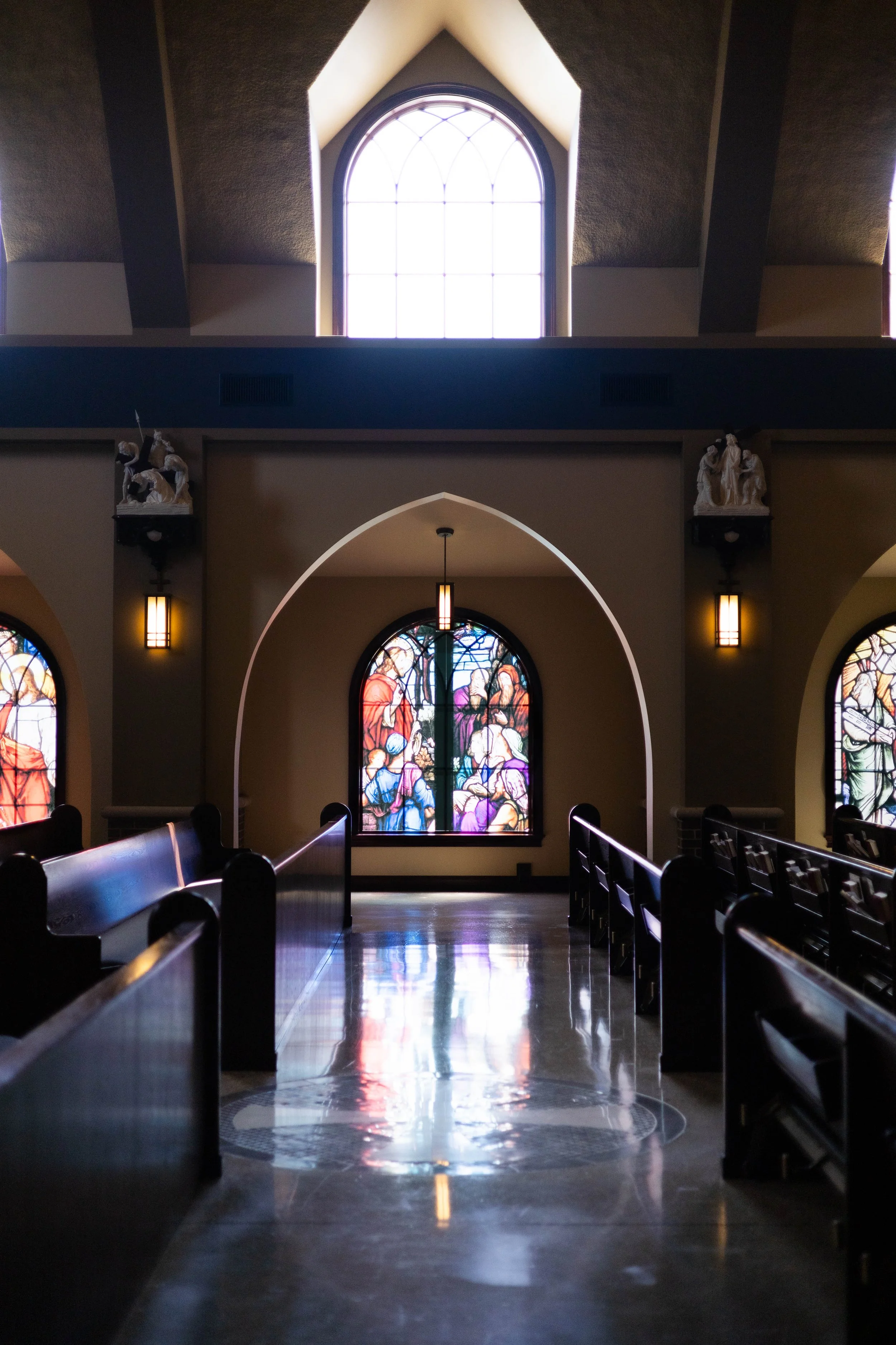 Interior view of a church aisle with dark wooden pews, stained glass windows, and a large arched window at the front, with church statues on either side of the window and pendant lights hanging from the ceiling.