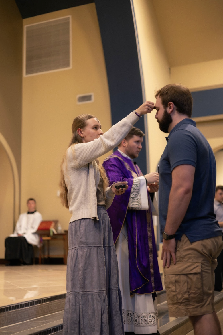 A woman applying a religious mark or blessing on a man's forehead during a church ceremony. A priest is in the background, and other attendees are present.
