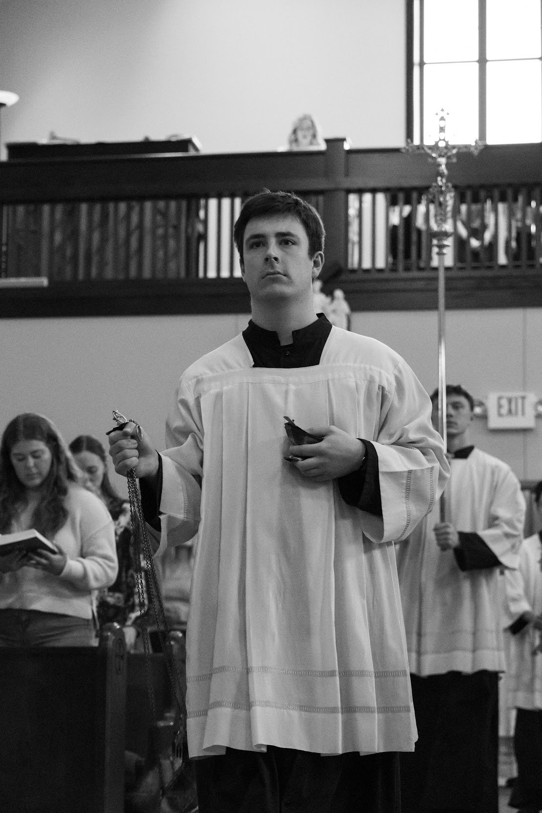 A young man in a religious choir robe holding a rosary and a black book standing inside a church with other choir members and a candelabrum in the background