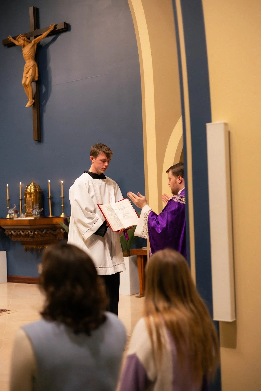 A Catholic mass ceremony showing a young man in a white robe and a priest in purple robes, reading from a religious book, with Catholic altar items and crucifix in the background, and congregation members seated in the foreground.