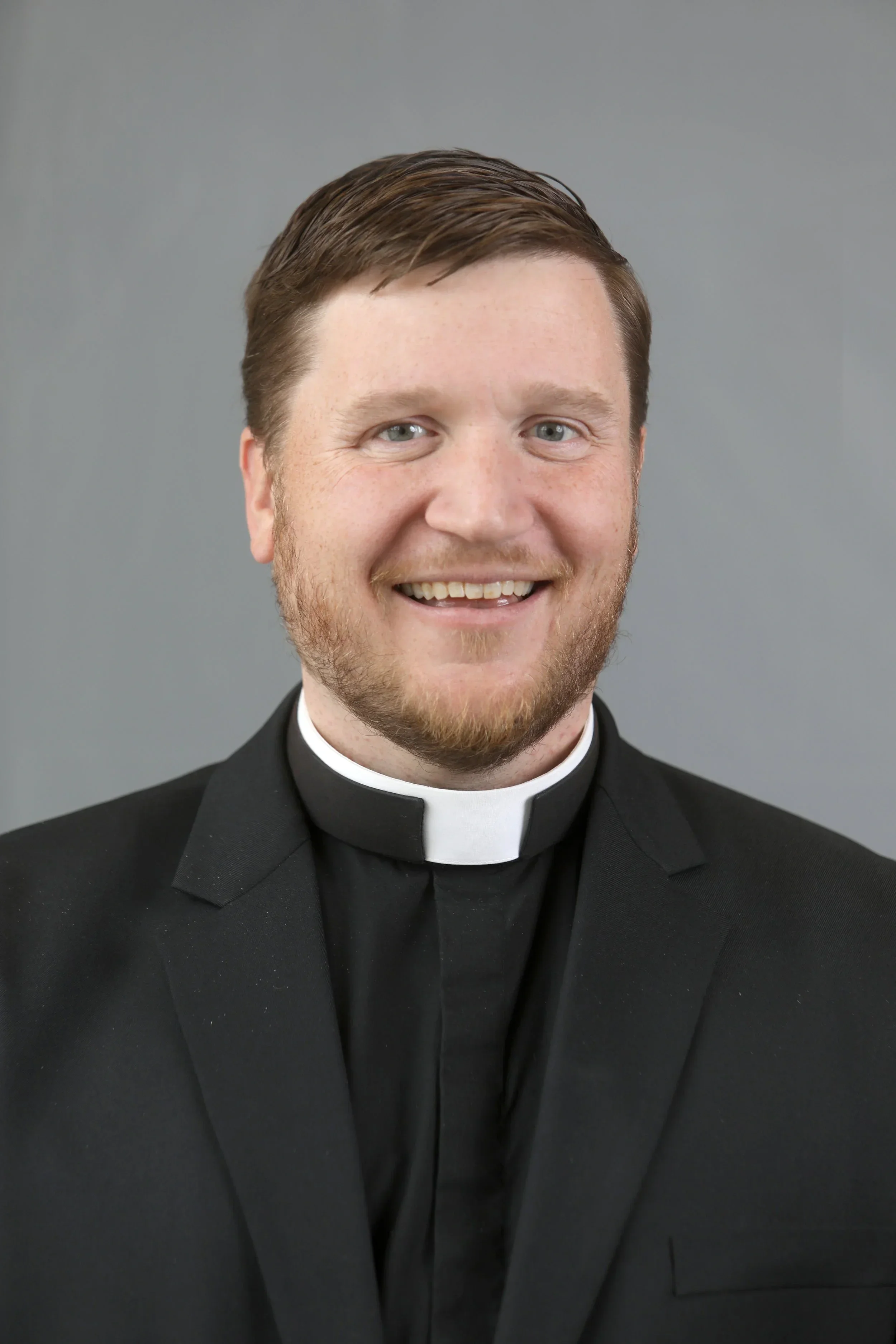 Close-up portrait of a smiling man in clerical attire, with a clerical collar and black suit, against a plain gray background.