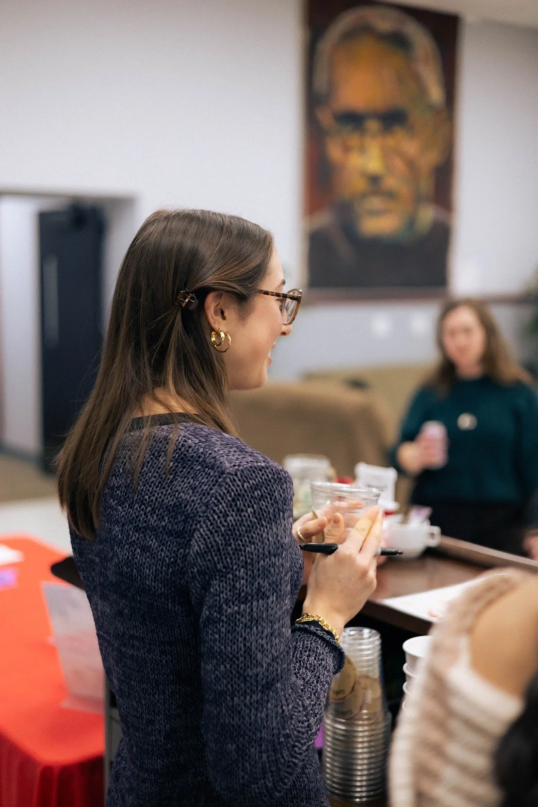 A woman with brown hair and glasses holding a clear cup and a spoon, standing at a table with cups and food, smiling and talking to someone off-camera. In the background, another woman is holding a smartphone, and a large abstract portrait painting hangs on the wall.