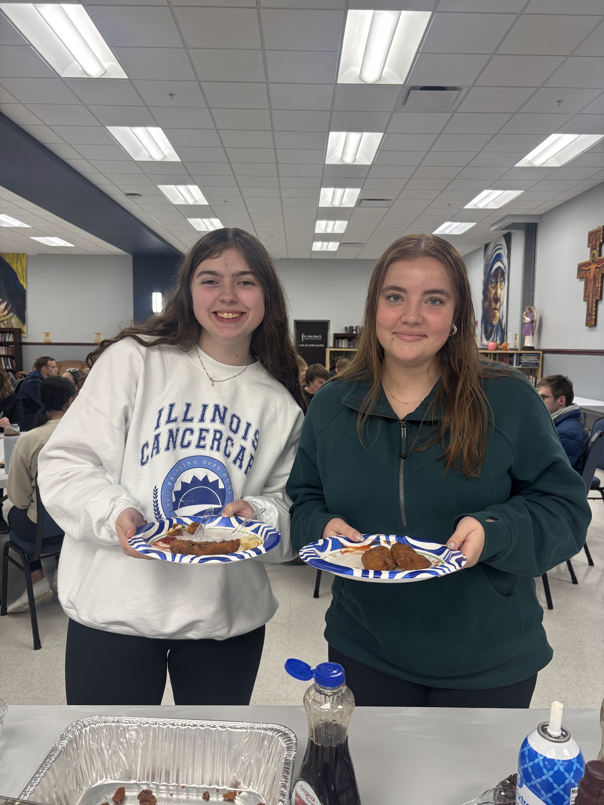 Two young women standing indoors, holding plates of food, smiling at the camera. The woman on the left wears a white sweatshirt with 'Illinois Cancer' written on it, and the woman on the right wears a dark green jacket. In the background, there are o