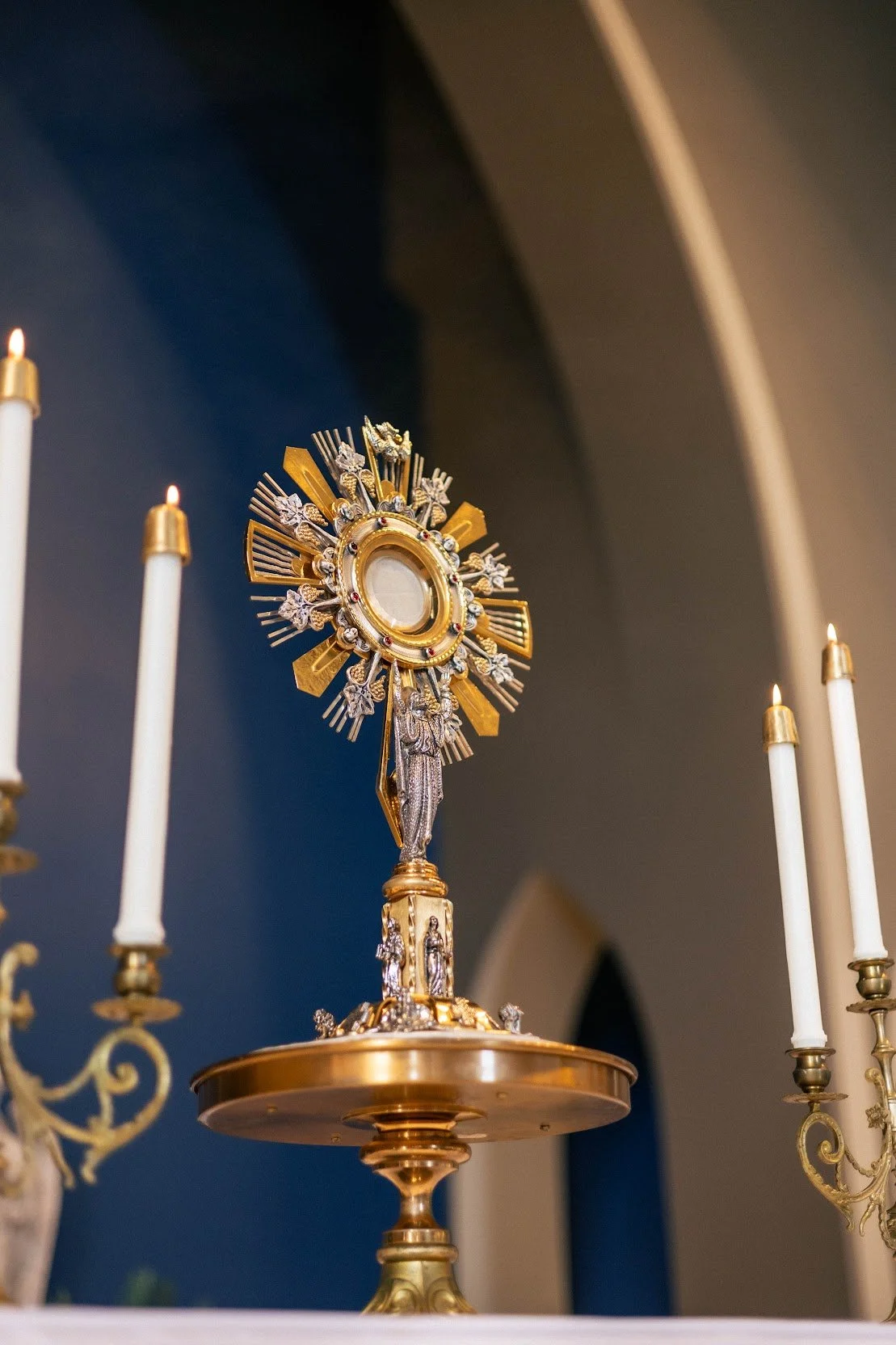Gold and silver monstrance with a circular center surrounded by rays, standing on a gold stand, surrounded by white candles in gold holders