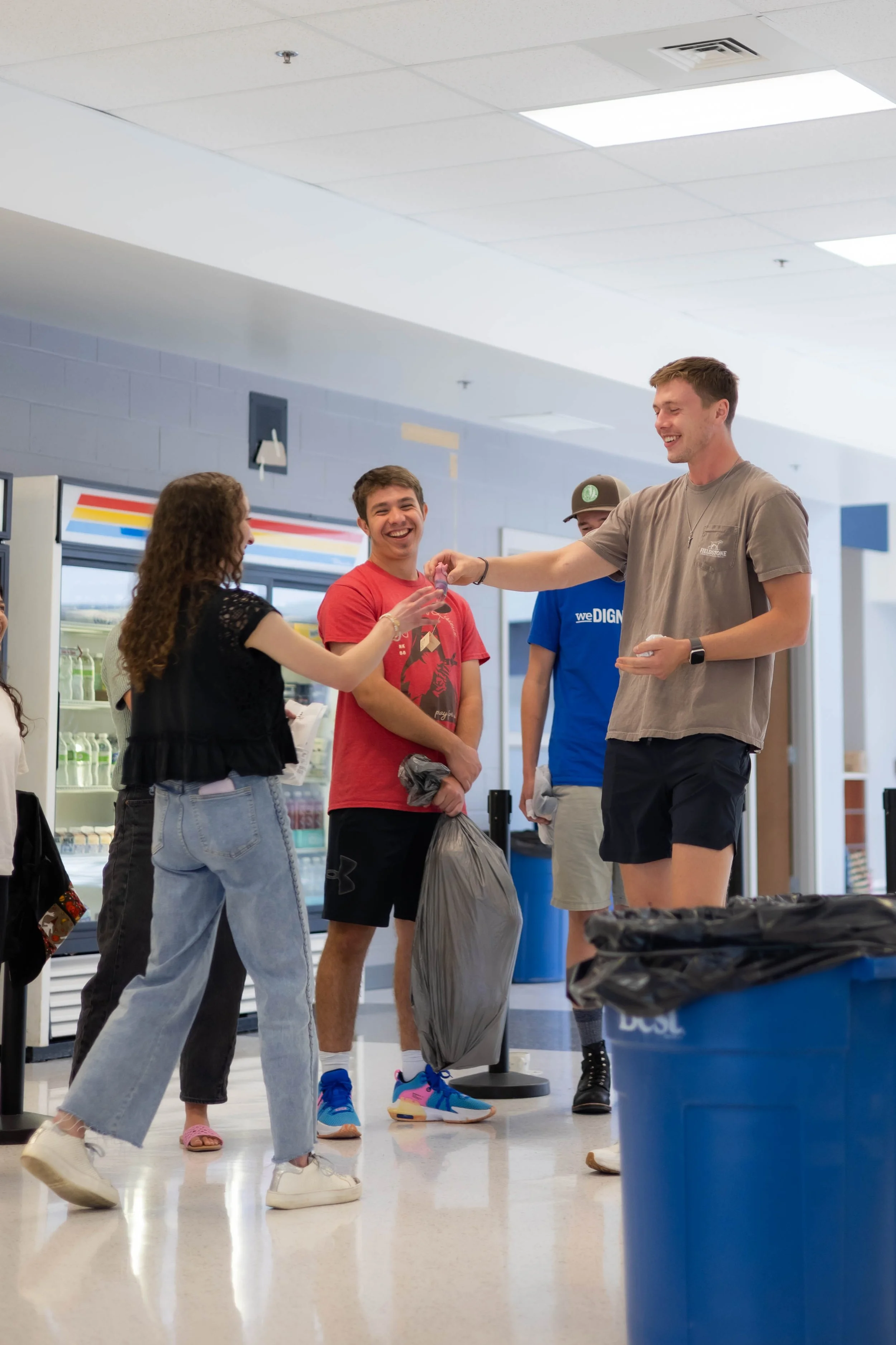 Group of young people inside a store, three of them are smiling and talking, one person is receiving a gift card, a person is holding a trash bag, and another is standing nearby. The store has refrigerators and a blue trash bin visible.