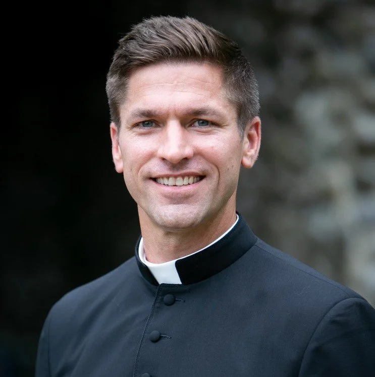 Portrait of a smiling man dressed as a priest, wearing a black clerical shirt with a white collar, outdoors with a blurred natural background.
