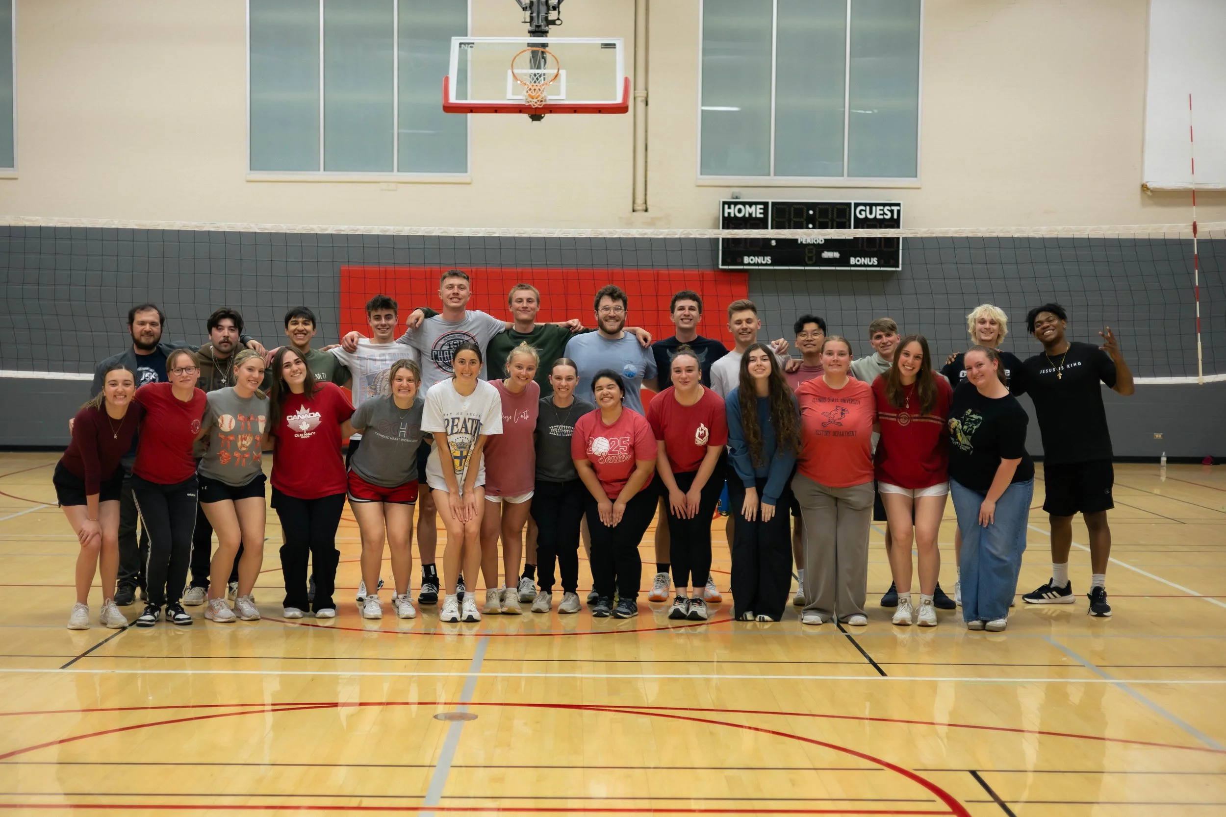 A large group of people, both men and women, standing together in a gymnasium with a volleyball net in front of them, smiling and posing for a photo.