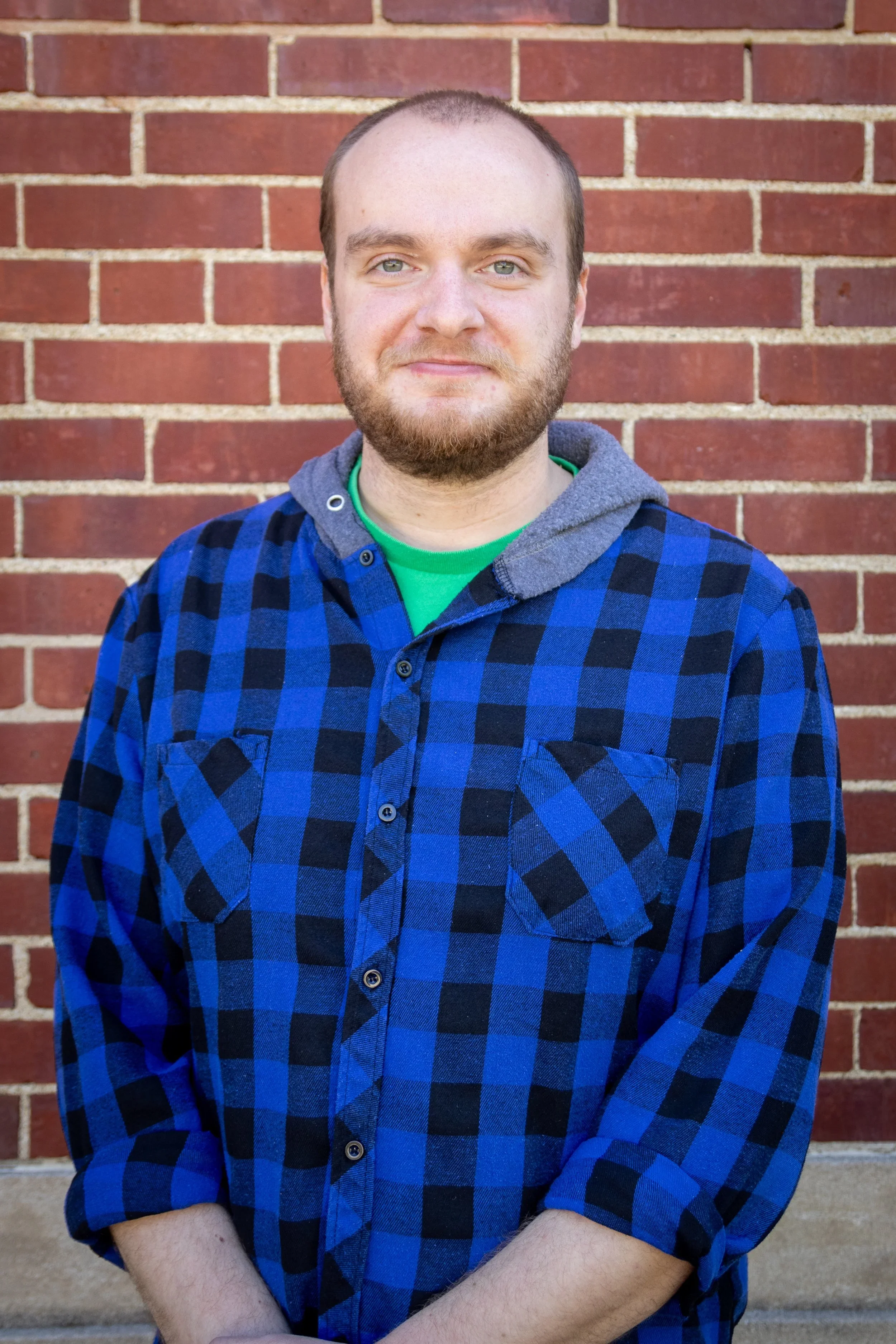 A young man with a beard and short hair standing in front of a brick wall. He is wearing a blue and black checkered shirt over a gray hoodie with a green shirt underneath. His arms are crossed in front of him.