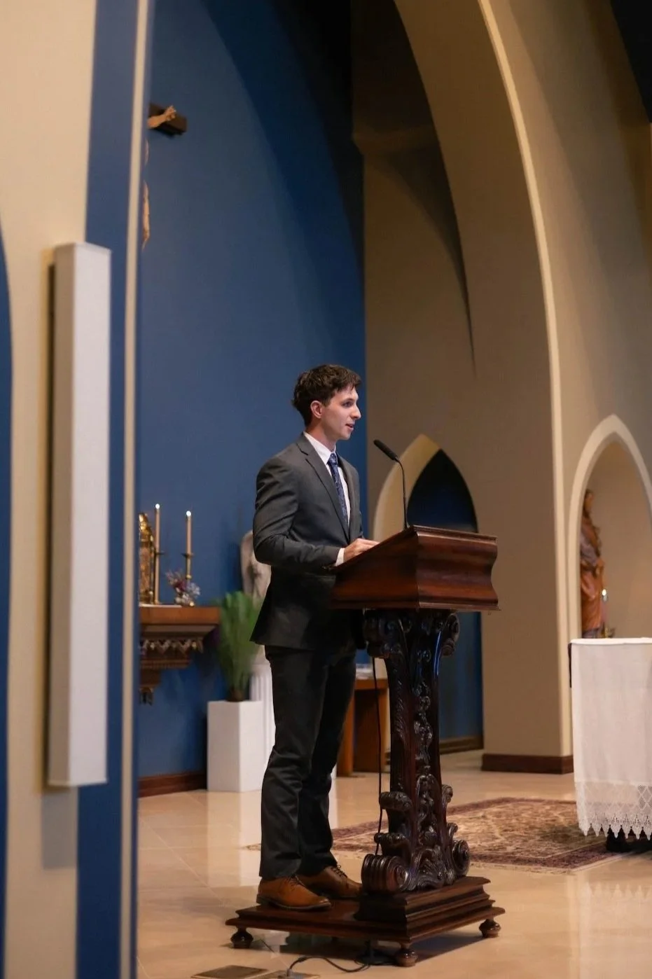 A young man in a gray suit is standing at a wooden pulpit with a microphone, delivering a speech or reading in a church with arched architecture and religious statues.