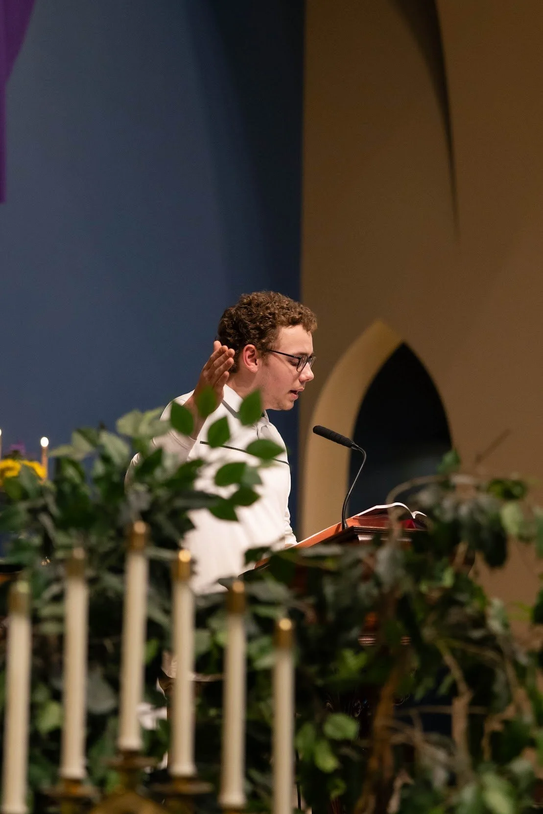 A young man with curly hair and glasses, dressed in a white shirt, stands at a podium with a microphone, raising his right hand and reading from a book or notes, in a church or similar setting decorated with green plants and candles.