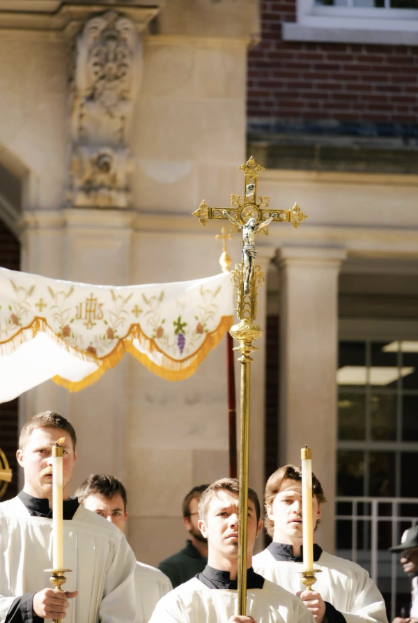 A religious procession with young men holding candles and a gold crucifix, taking place outdoors near a church building.