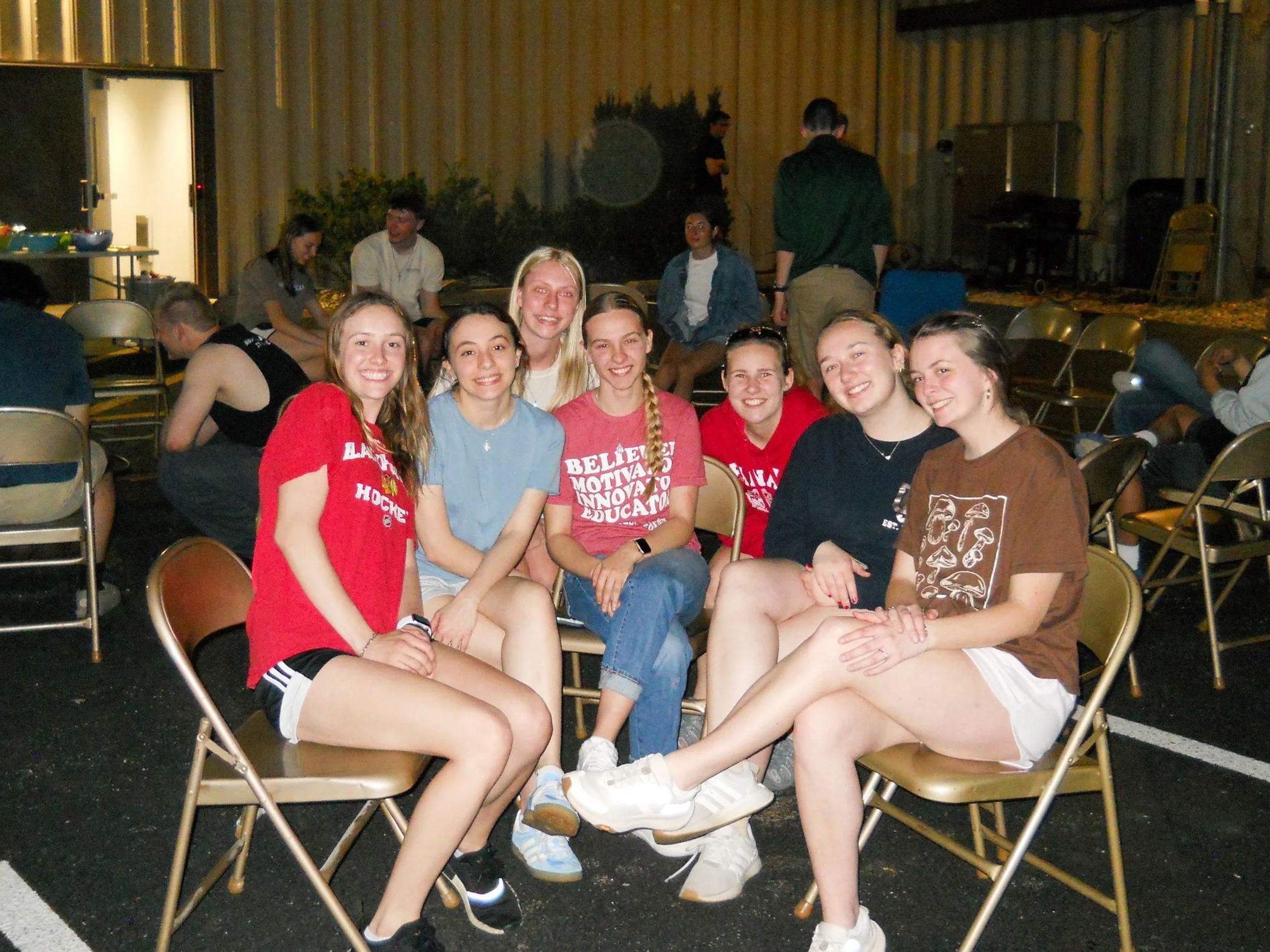 Group of young women sitting on chairs and smiling, with other people in the background, outside at bonfire event.