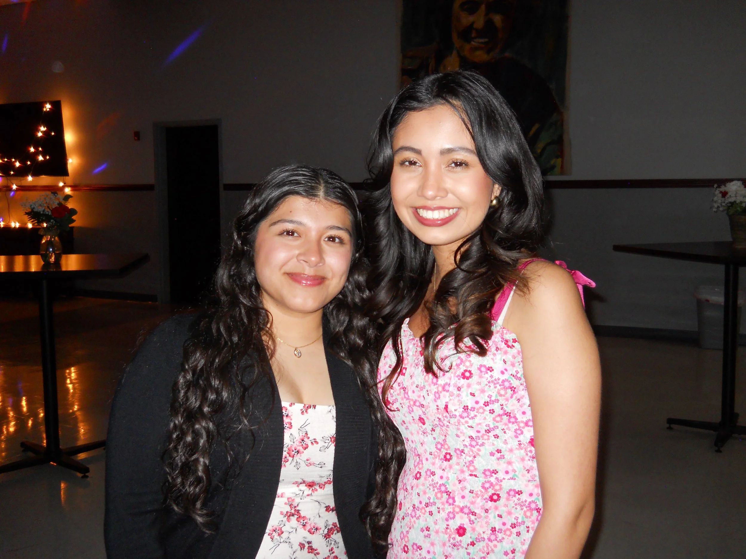 Two young women with long, dark hair posing together indoors at a social event. One is wearing a black cardigan over a floral dress, and the other is in a pink floral sleeveless dress. There are decorative lights and a large painting in the backgroun