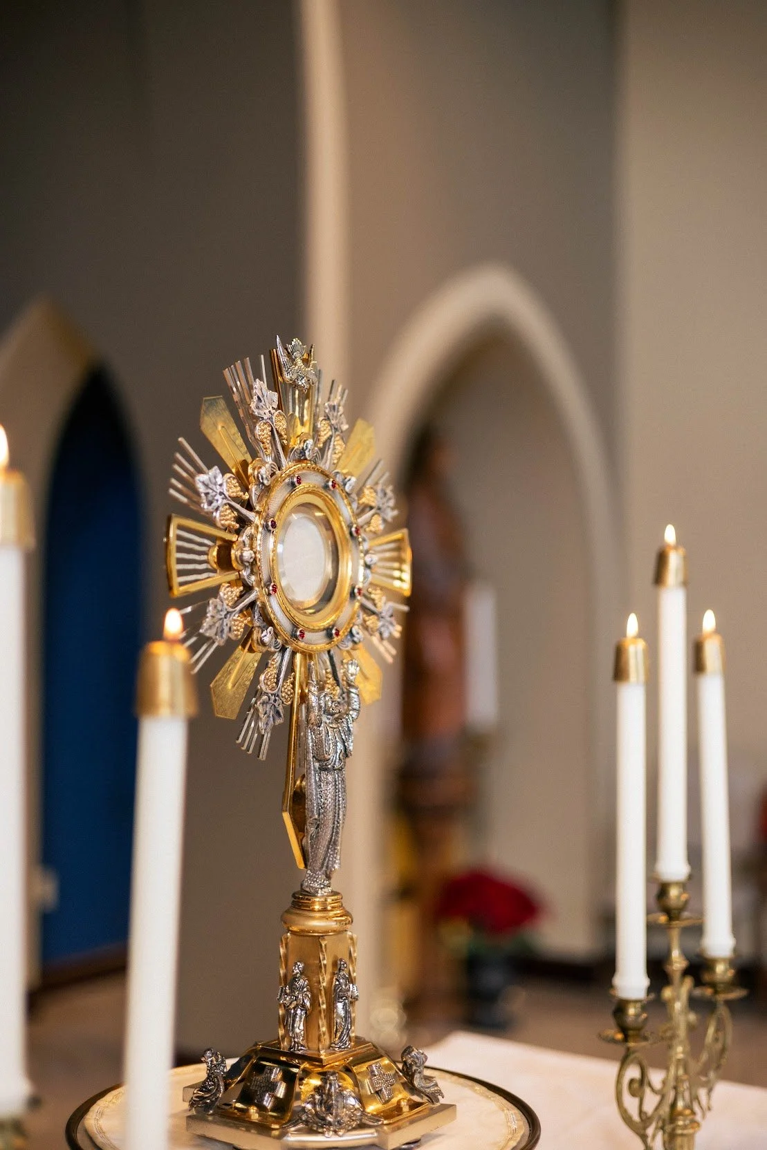 Gold and silver religious monstrance with a circular center, surrounded by rays and decorative figures, on a table with candles.