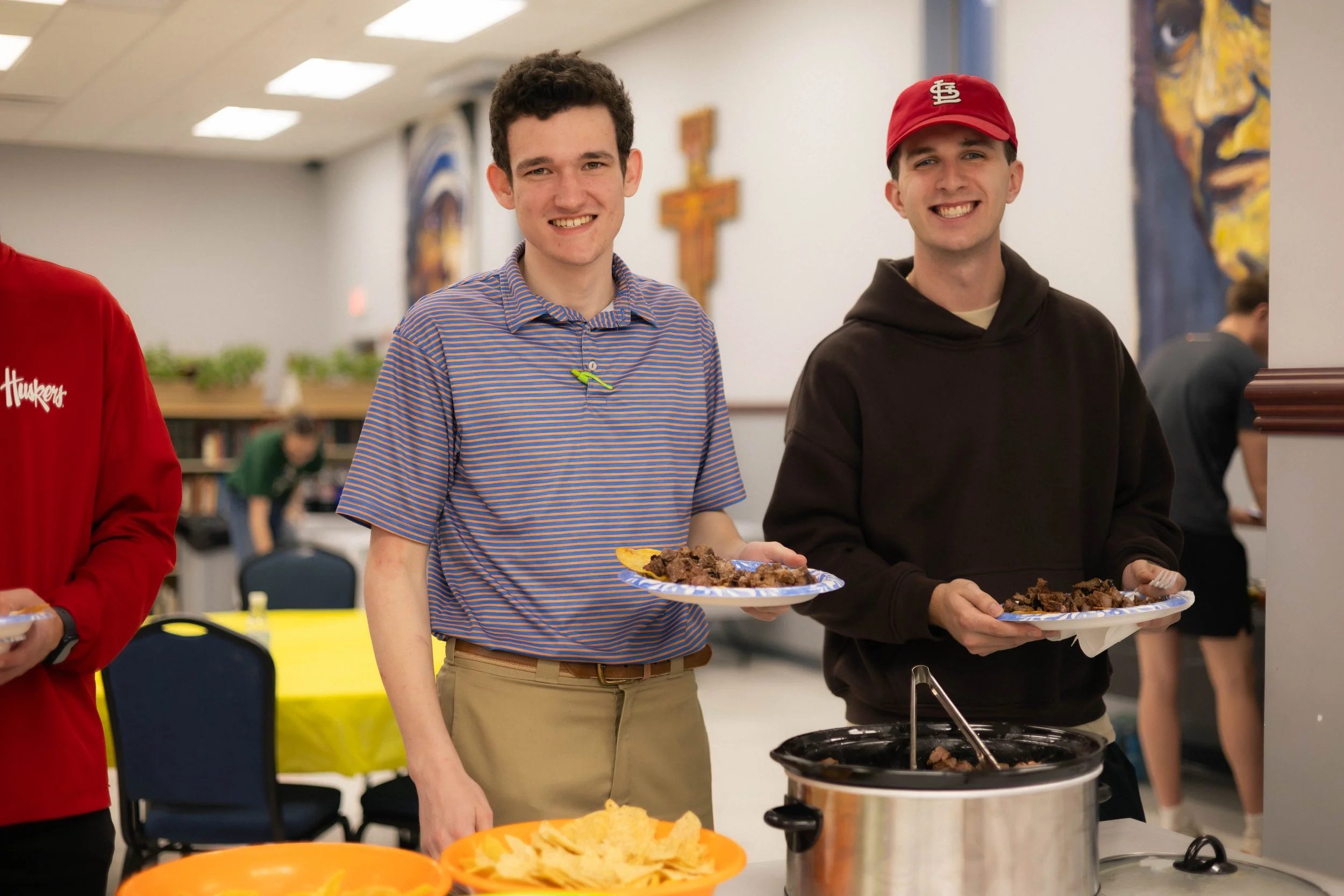 Two young men smiling while holding plates of food at a buffet or potluck event. The background shows a community setting with tables, chairs, and artwork on the walls.