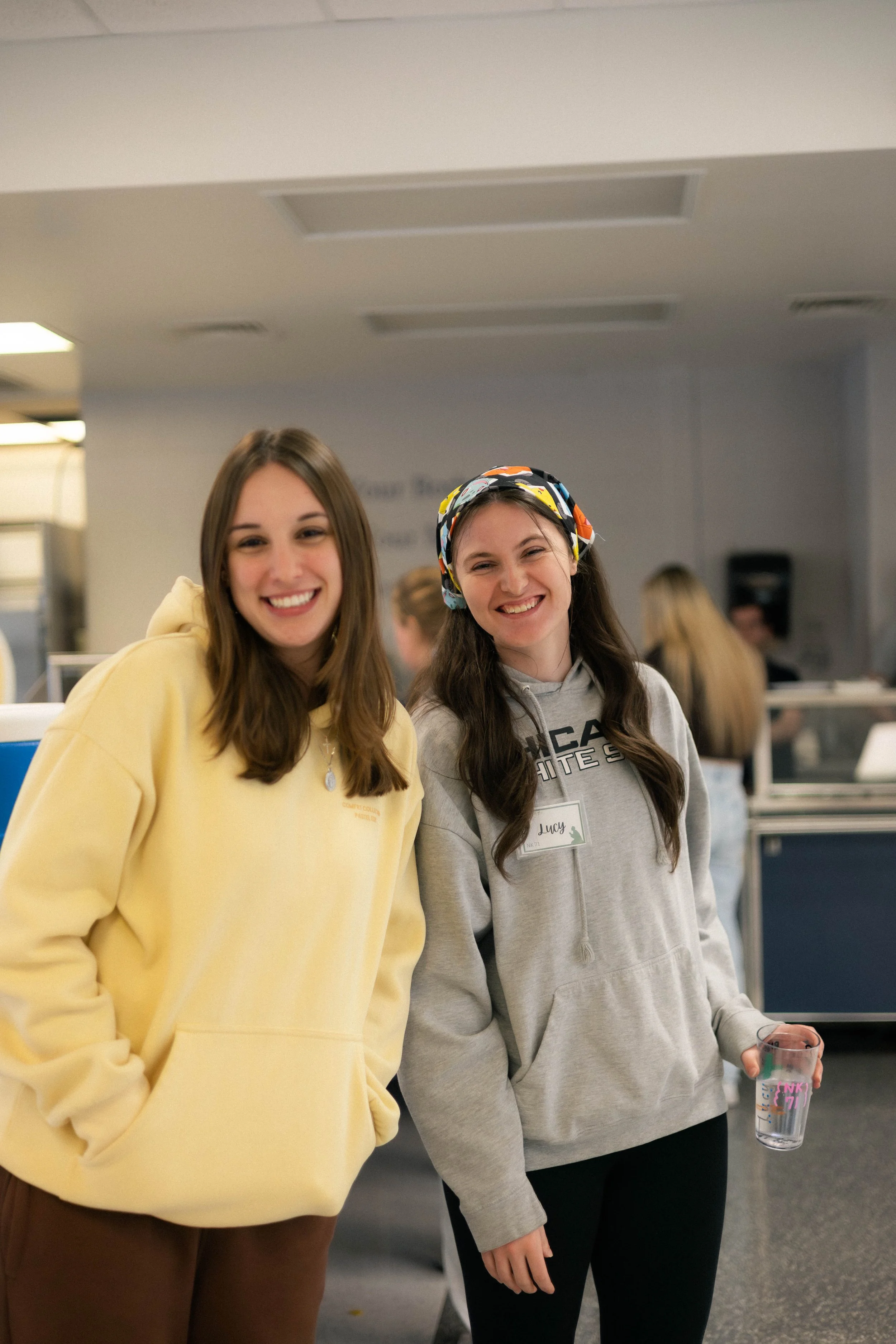 Two young women smiling at the camera in an indoor setting, possibly a school or workplace. One wears a yellow hoodie, the other a gray hoodie with a name tag that says "Lucy" and a colorful headband. The woman on the right is holding a transparent g