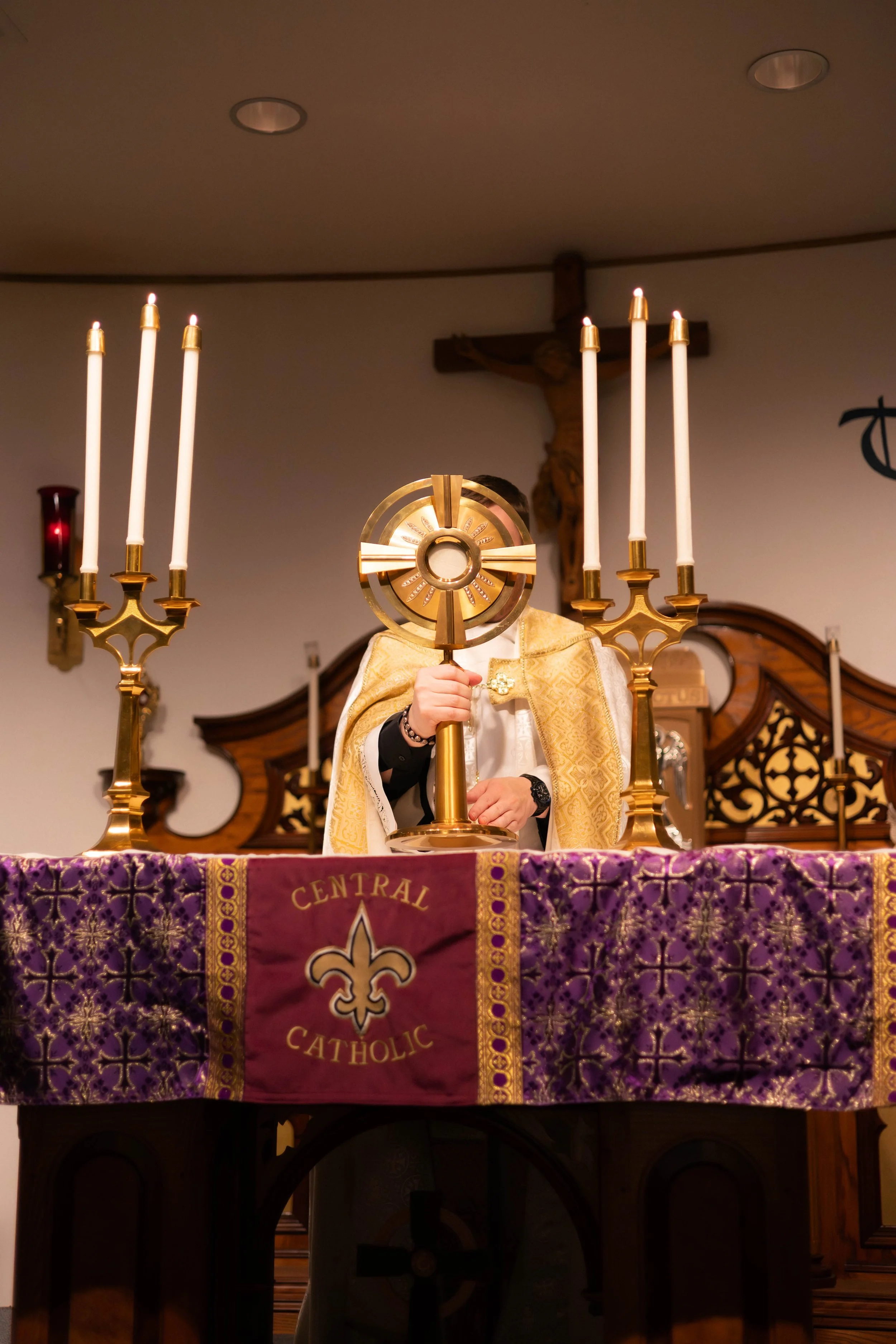 A priest holding a monstrance during a Catholic Holy Eucharist ceremony in front of a crucifix, with a purple altar cloth with gold embroidery and four lit candles on gold candlesticks.