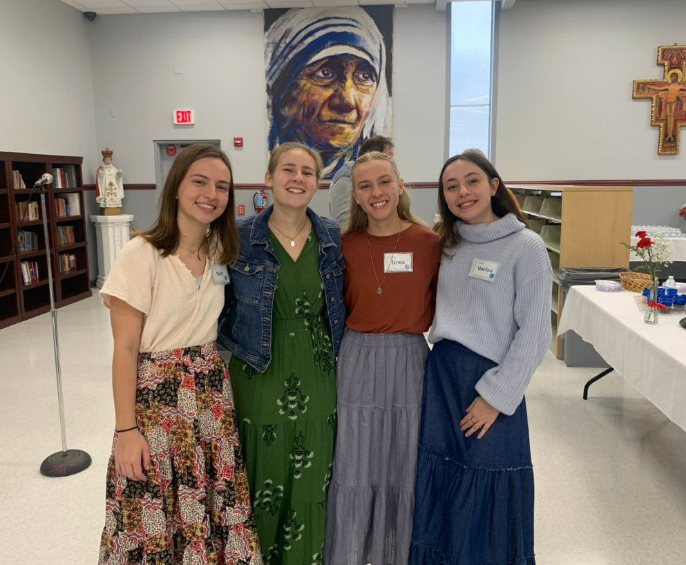 Four young women standing together indoors, smiling at the camera. Behind them is a colorful mural of a woman with a headscarf, and to the right, a table with flowers and other items.