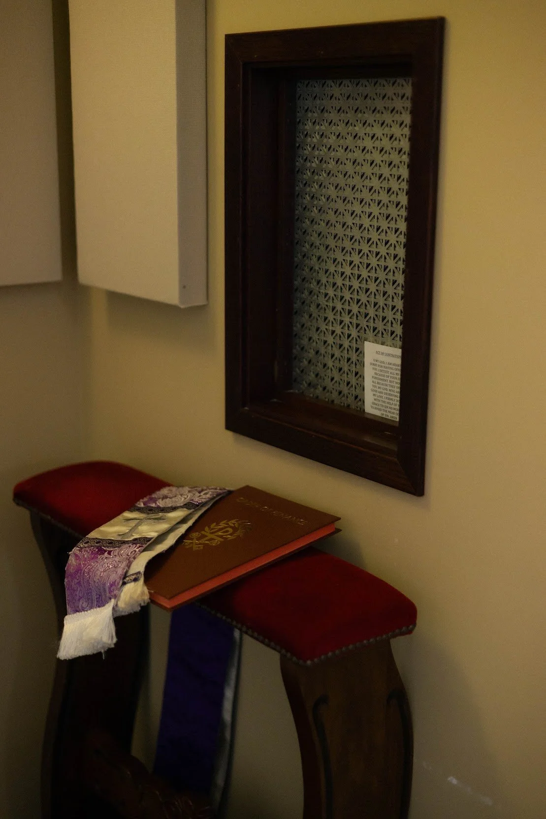 A hallway corner with a wall-mounted decorative mirror with a dark wooden frame, a red cushioned stool, and a small table with books and a decorative scarf. There are also white cabinets mounted on the wall.