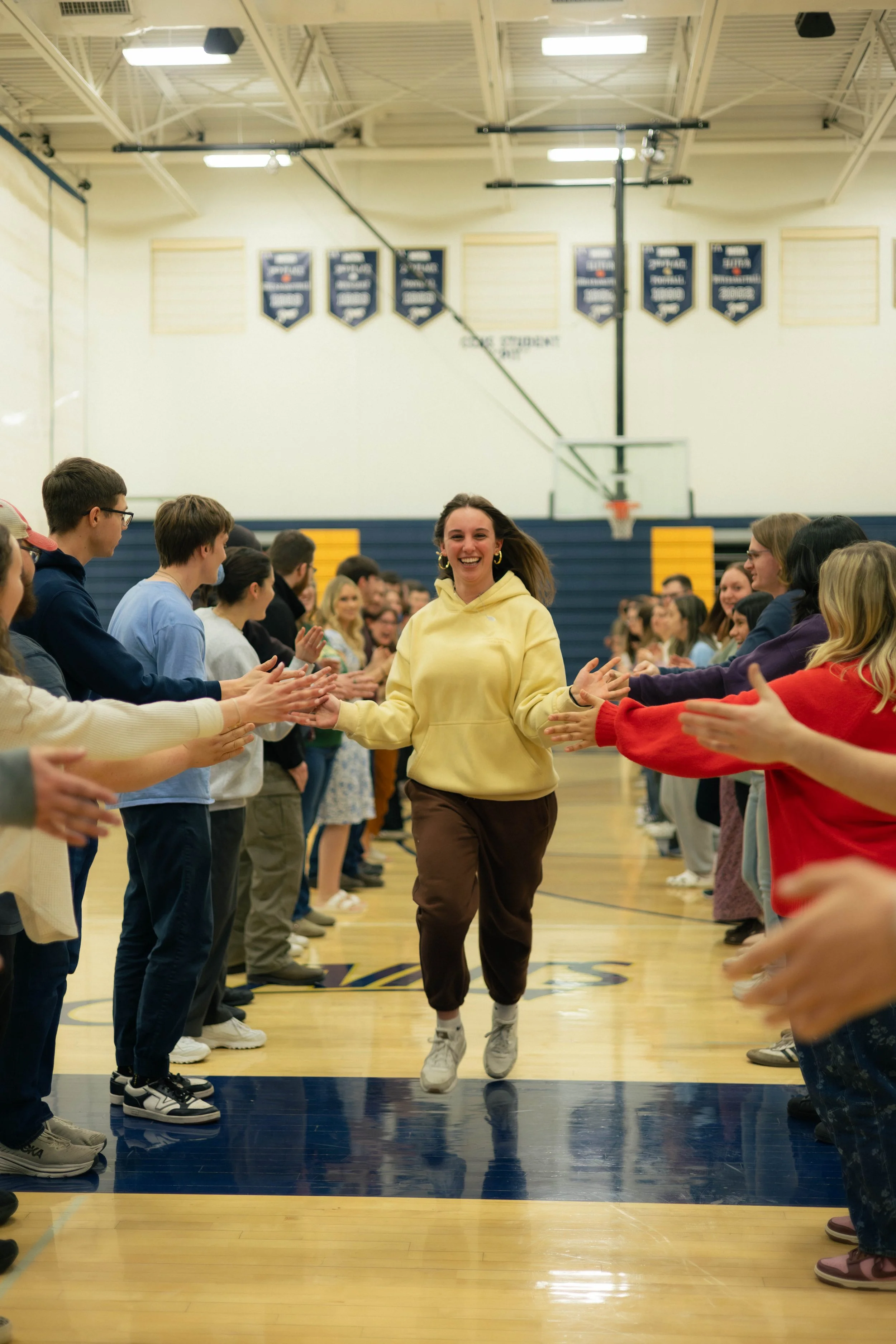 A young woman in a yellow hoodie and brown pants is running down basketball court, smiling, while people on both sides clap and cheer for her.