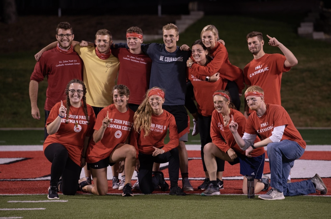Group of young athletes celebrating on a football field at night, wearing red and yellow shirts with school names and slogans, some with face paint and headbands, standing and kneeling in a group pose.